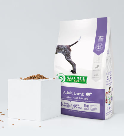 Ein Beutel Nature's Protection NP Lamm & Reis 18 kg Breeder Bag steht aufrecht neben einem weißen Würfel, auf dem und um den herum Krokantstücke verstreut sind. Auf der Verpackung sind ein Teil eines Hundes und Produktangaben abgebildet.